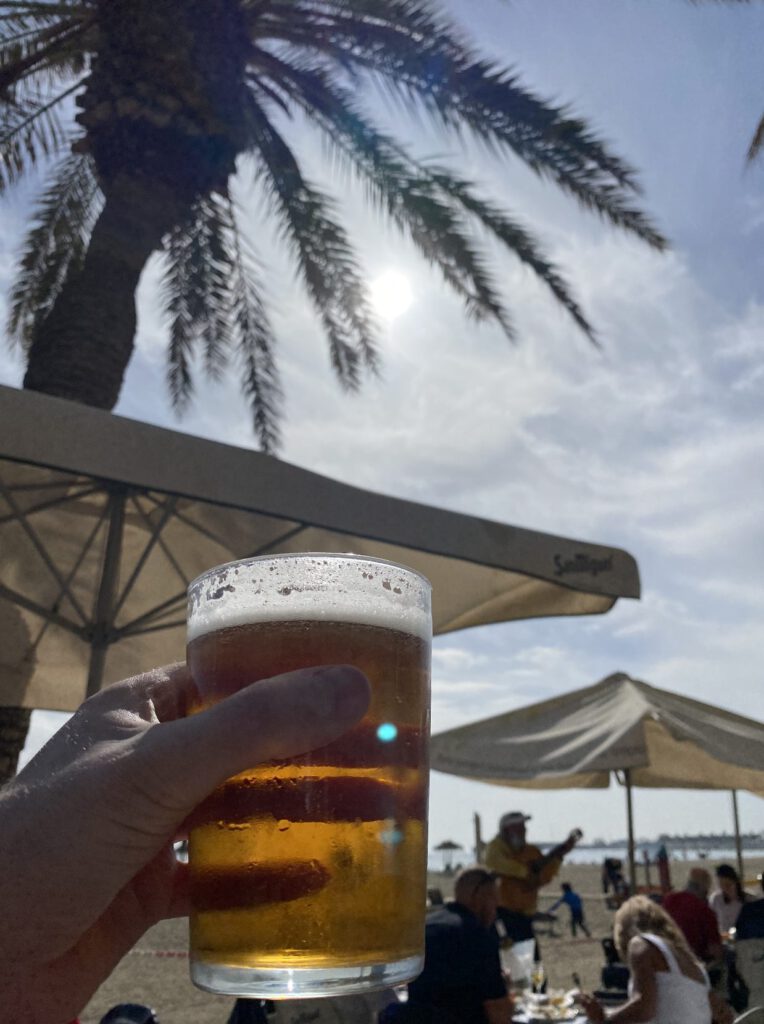 Hand met een koud biertje onder een strandparasol en palmboom in de zon, met een gitarist op de achtergrond